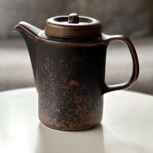 May include: A brown ceramic teapot with a lid and handle. The teapot has a textured surface with a speckled pattern. The spout is on the left side, and the handle is on the right. The lid has a small knob on top.