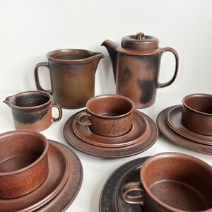 May include: A set of brown ceramic tableware, including a teapot with a lid, two jugs, cups, and saucers. The pieces have a speckled, textured appearance with dark brown and black accents. The set is arranged on a white surface.