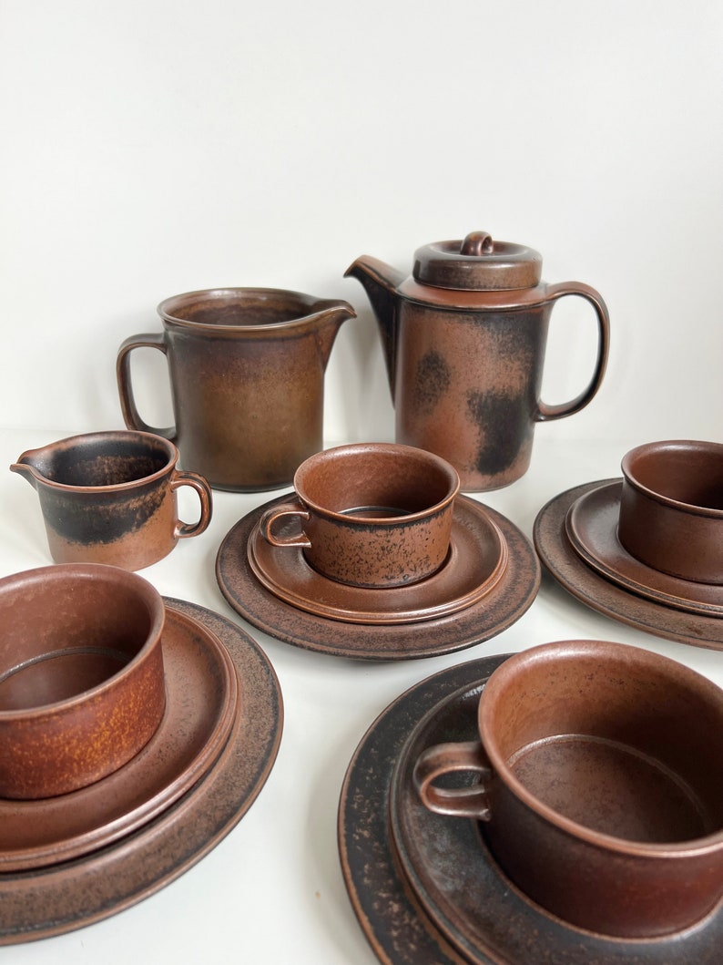 May include: A set of brown ceramic tableware, including a teapot with a lid, two jugs, cups, and saucers. The pieces have a speckled, textured appearance with dark brown and black accents. The set is arranged on a white surface.