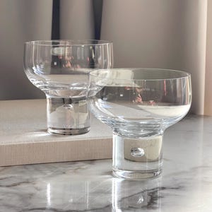 May include: Two clear, stemless wine glasses with a rounded bowl and a short, cylindrical stem. The glasses sit on a marble surface, reflecting light. The design features a small, bubble-like detail in the stem.