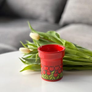 May include: A red ceramic flower pot with a tulip pattern. The pot is filled with white tulips and green leaves.