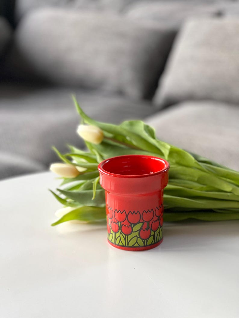 May include: A red ceramic flower pot with a tulip pattern. The pot is filled with white tulips and green leaves.