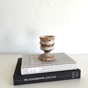 May include: Two books stacked on top of each other. The top book is a white and grey patterned book with the title "The Little Women" by Louisa May Alcott. The bottom book is black with the title "The Handbook of Style" and the text "Esquire Heart Books". A small, white ceramic vase with brown speckles sits on top of the books.