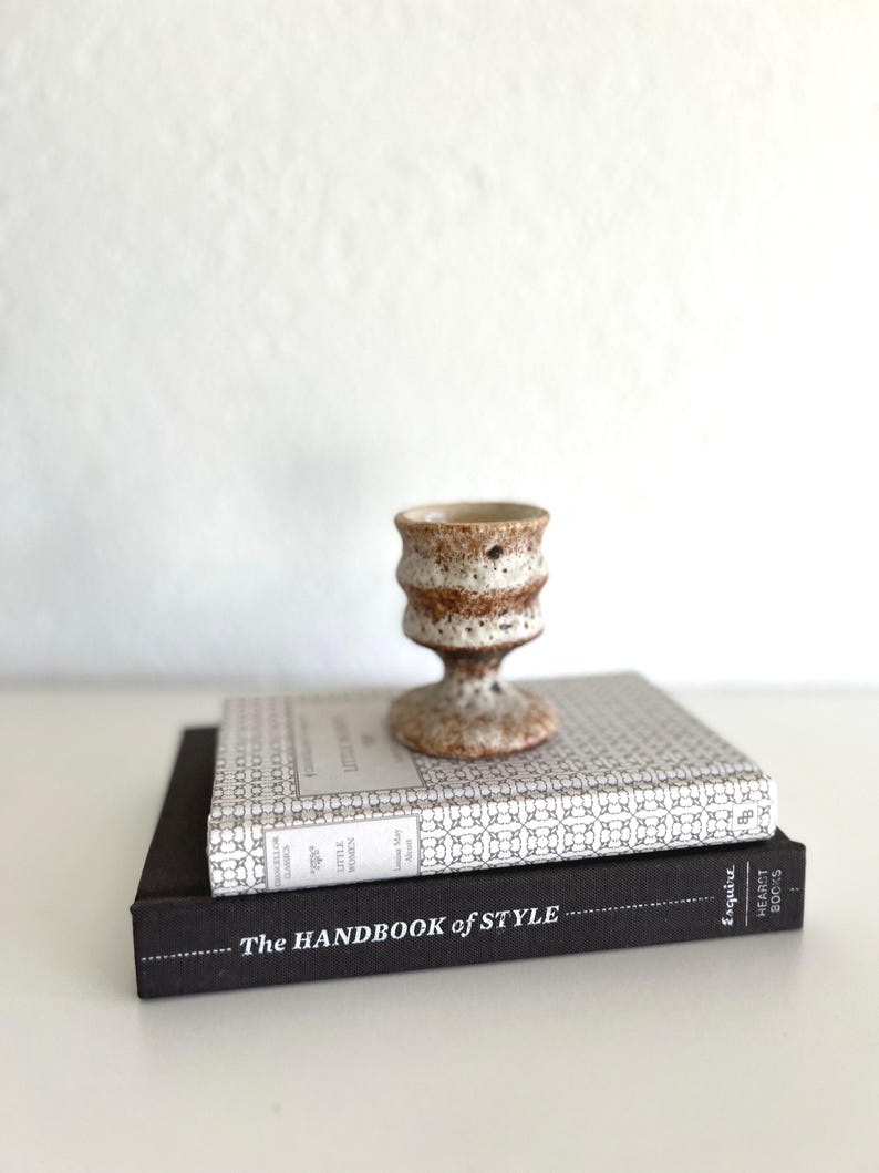May include: Two books stacked on top of each other. The top book is a white and grey patterned book with the title "The Little Women" by Louisa May Alcott. The bottom book is black with the title "The Handbook of Style" and the text "Esquire Heart Books". A small, white ceramic vase with brown speckles sits on top of the books.