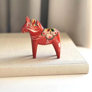 May include: A small, red wooden Dala horse figurine with white and yellow painted details. The horse has a saddle design with floral patterns and is standing on a neutral-coloured surface. A classic Scandinavian folk art piece.