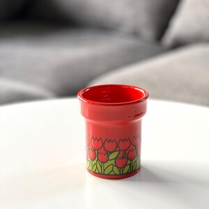 May include: A red ceramic cup with a tulip pattern. The cup is about 4 inches tall and has a diameter of about 3 inches.