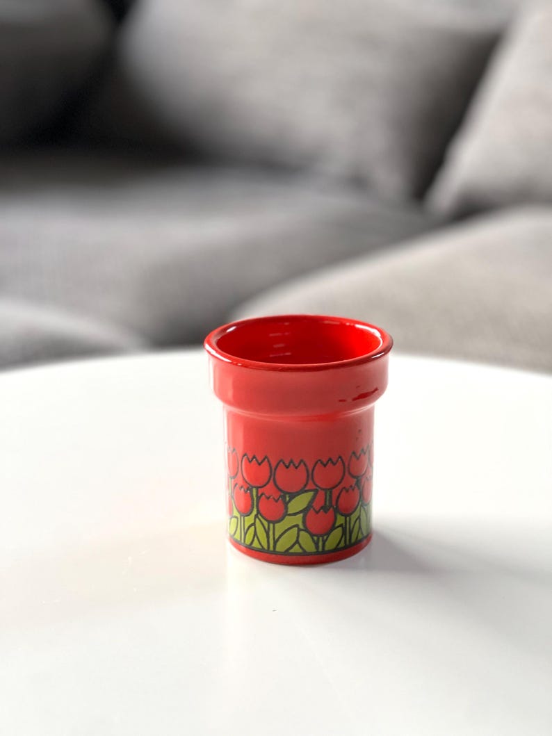 May include: A red ceramic cup with a tulip pattern. The cup is about 4 inches tall and has a diameter of about 3 inches.