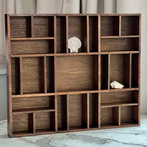 Op de afbeelding: Een houten display plank met meerdere compartimenten van verschillende afmetingen. De plank is gebeitst in een warme bruine kleur en heeft twee schelpen. De plank is ontworpen om kleine items en verzamelobjecten in op te bergen.