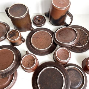 May include: A collection of brown ceramic tableware, including plates, saucers, cups, and a teapot. The pieces have a speckled texture and a white rim. The set is arranged on a white surface, showcasing a complete dining set.