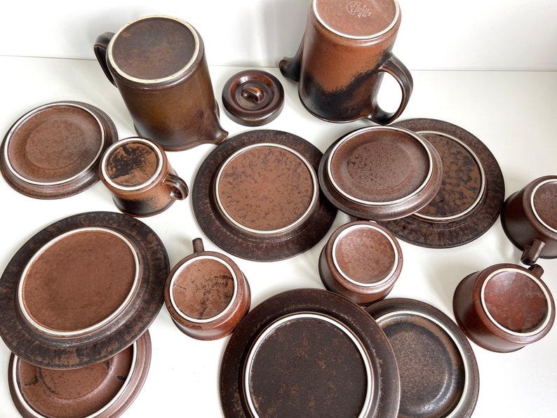 May include: A collection of brown ceramic tableware, including plates, saucers, cups, and a teapot. The pieces have a speckled texture and a white rim. The set is arranged on a white surface, showcasing a complete dining set.