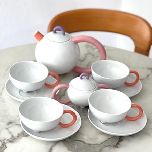 May include: A white ceramic tea set with a teapot, sugar bowl, and four teacups on saucers. The teapot has a pink handle and orange spout. The teacups have orange handles. The sugar bowl and teapot have purple handles. A modern tea set.