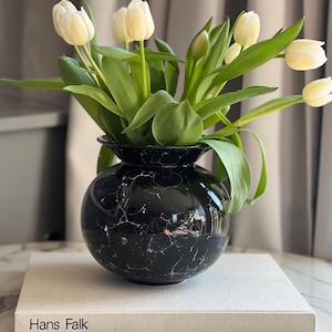 Può includere: Un vaso nero effetto marmo pieno di tulipani bianchi. Il vaso è posto su un libro con il titolo "Hans Falk". I tulipani hanno steli e foglie verdi, creando contrasto con il vaso scuro. Elegante composizione floreale.