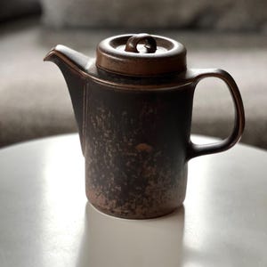 May include: A brown ceramic teapot with a lid and handle. The teapot has a textured surface and a spout for pouring. The lid has a small loop handle. The teapot is sitting on a white surface.
