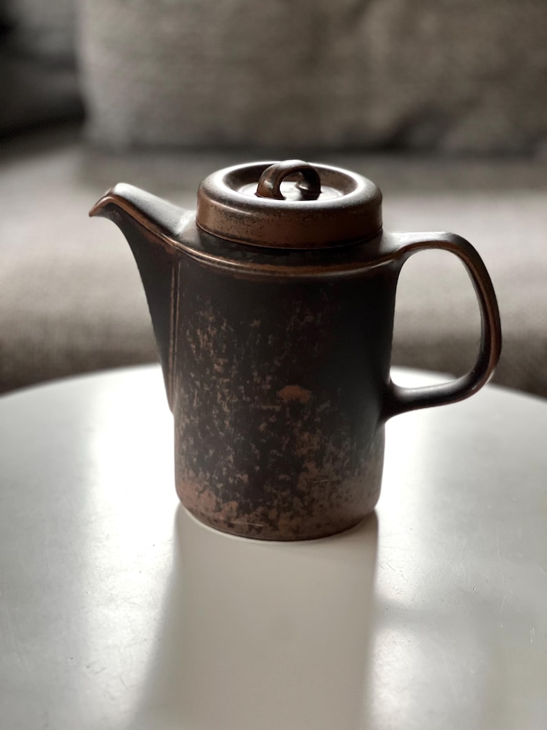 May include: A brown ceramic teapot with a lid and handle. The teapot has a textured surface and a spout for pouring. The lid has a small loop handle. The teapot is sitting on a white surface.