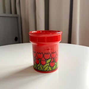 May include: A red ceramic cup with a tulip pattern. The cup has a white background with green leaves and red tulips.