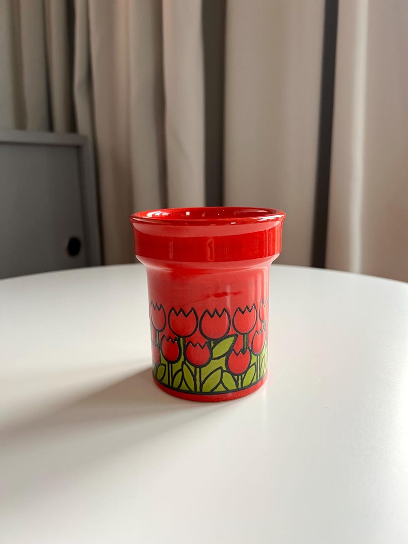 May include: A red ceramic cup with a tulip pattern. The cup has a white background with green leaves and red tulips.