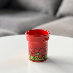 May include: Red ceramic cup with a tulip pattern. The cup has a wide rim and a cylindrical shape.