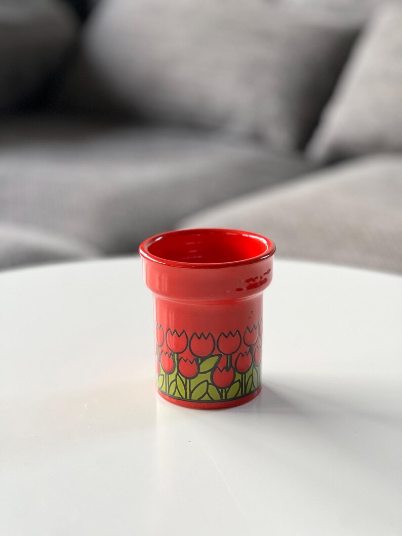 May include: Red ceramic cup with a tulip pattern. The cup has a wide rim and a cylindrical shape.