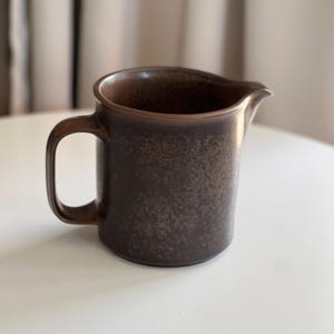 May include: A brown ceramic jug with a handle and a spout. The jug has a speckled texture and a dark brown colour. The handle is rectangular and attached to the side of the jug.