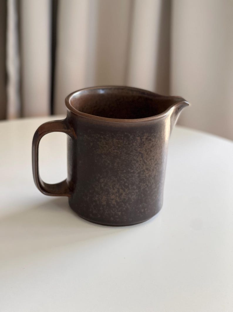 May include: A brown ceramic jug with a handle and a spout. The jug has a speckled texture and a dark brown colour. The handle is rectangular and attached to the side of the jug.