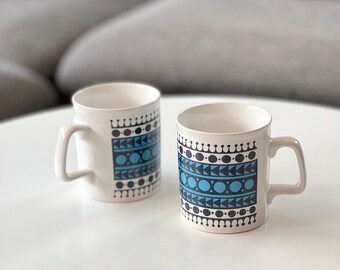 May include: Two white ceramic mugs with a blue and black geometric pattern. Each mug has a handle and a cylindrical shape. The mugs are on a white surface, with a gray sofa in the background.
