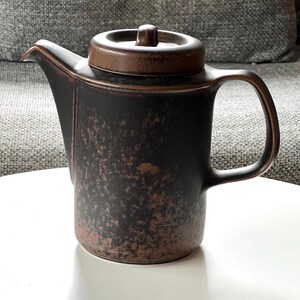 May include: A dark brown ceramic teapot with a matching lid. The teapot has a handle and a spout. The surface has a textured, mottled appearance. The teapot is sitting on a white surface.