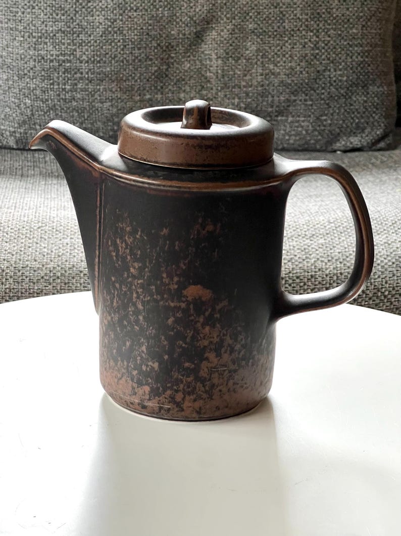 May include: A dark brown ceramic teapot with a matching lid. The teapot has a handle and a spout. The surface has a textured, mottled appearance. The teapot is sitting on a white surface.