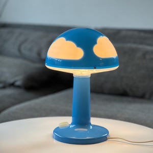 May include: A blue mushroom-shaped table lamp with a white base and stem. The lamp's shade is blue with two white cloud cutouts that illuminate when the lamp is on. The lamp is on a white table, with a gray sofa in the background.