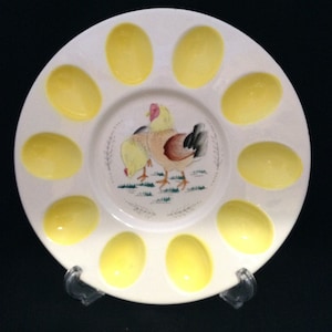 May include: A white ceramic egg plate with 12 yellow egg-shaped indentations. The center of the plate features a painted image of two chickens.