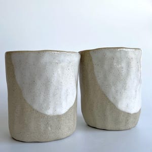 May include: Two handmade ceramic cups. Each cup has a beige base and a white, speckled glaze that covers the top half. The cups have a simple, modern design, ideal for coffee or tea.