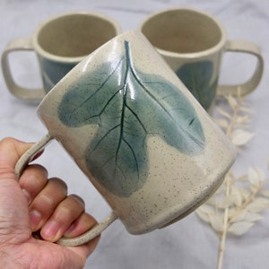 May include: A white ceramic mug with a blue leaf design. The mug has a speckled glaze and a handle.