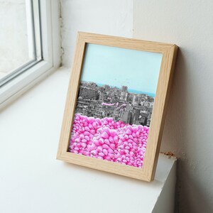 May include: A framed print featuring a cityscape in grayscale with a light blue sky and a foreground of vibrant pink flower-like shapes. The frame is made of light-colored wood, resting on a white surface.
