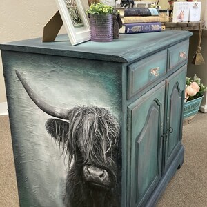May include: A teal blue painted cabinet with two drawers and two doors. The cabinet has a distressed finish and features a black and white highland cow illustration on the side.