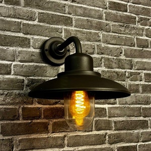 Puede incluir: Aplique de pared exterior negro con una pantalla redonda de metal negro y una bombilla de vidrio transparente. La luz está montada en una pared de ladrillo.