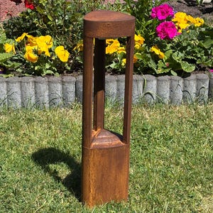 May include: A weathered, rectangular outdoor lamp post with a cylindrical top. The lamp post has a hollow, open design with vertical supports. The rusty brown metal contrasts with the green grass and colourful flowers in the background.