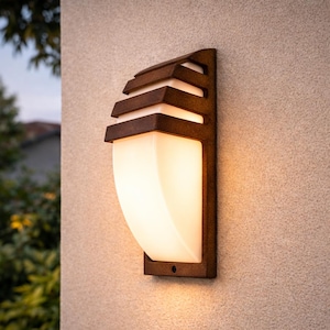 Puede incluir: Una lámpara de pared para exteriores de color bronce. La lámpara tiene una base rectangular con una pantalla de vidrio esmerilado curvada. La parte superior de la lámpara tiene un diseño en capas. La luz está montada en una pared texturizada de color claro.