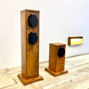 Peut inclure: Deux prises électriques extérieures en métal rouillé avec des prises noires. La plus grande prise a deux prises et la plus petite prise en a une. Les deux prises sont sur une surface en bois.