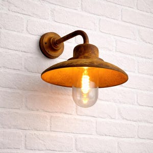 May include: A rustic, wall-mounted outdoor light fixture with a weathered, brown finish. The fixture features a curved arm, a circular backplate, and a downward-facing, bell-shaped shade. A clear glass bulb is visible, emitting a warm, yellow glow.