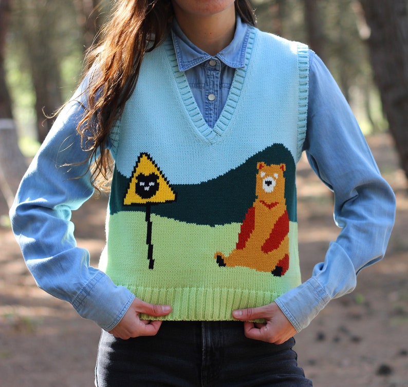 May include: A light blue knitted vest with a yellow triangle, a black cat, and a brown bear on a green and blue background.