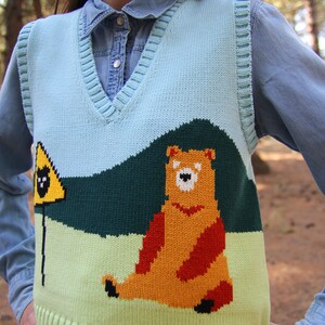 May include: A light blue knitted vest with a green, yellow, and orange bear design. The bear is sitting on a green hill with a yellow warning sign with a black bear inside.