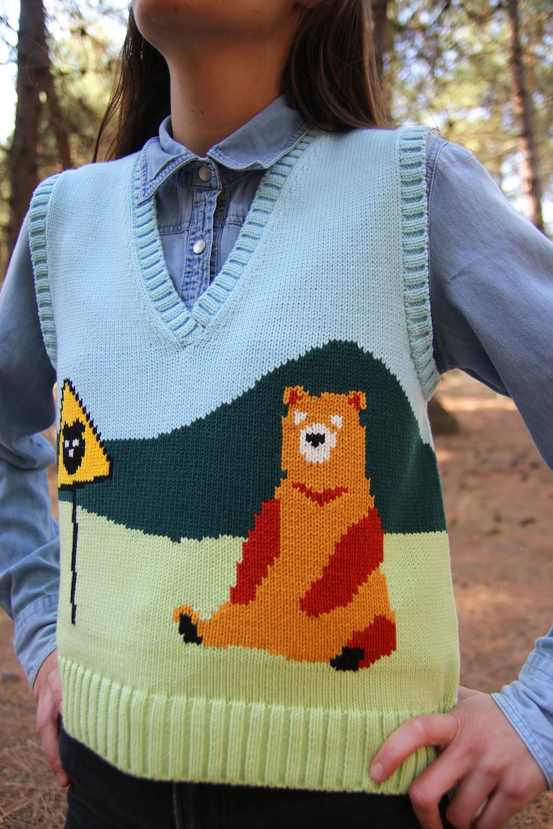 May include: A light blue knitted vest with a green, yellow, and orange bear design. The bear is sitting on a green hill with a yellow warning sign with a black bear inside.