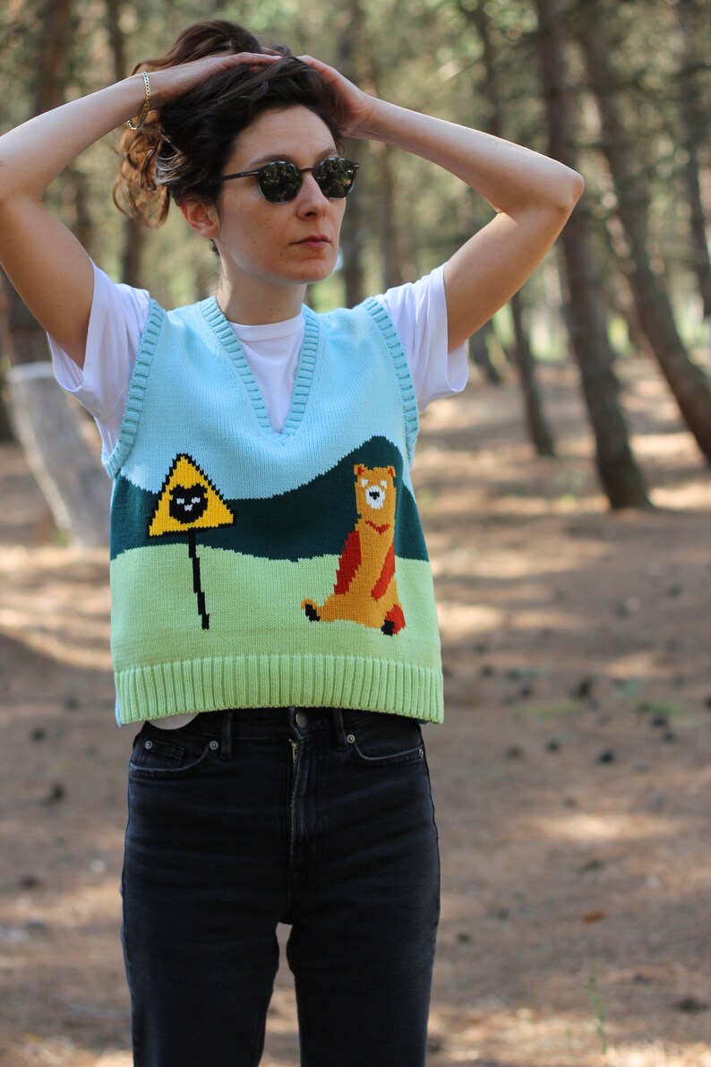 May include: A light blue knitted vest with a green, yellow, and orange bear and a black triangle with a black cat graphic. The vest has a V-neck and is worn over a white t-shirt.