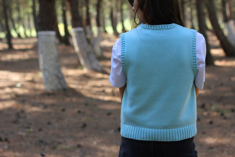 May include: A light blue knitted vest with a ribbed hem and a white t-shirt underneath.