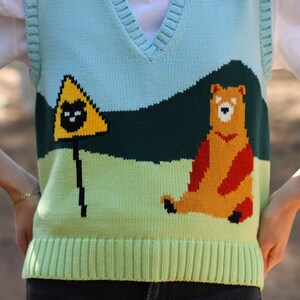 May include: A light blue, green, and yellow knitted vest with a bear and a warning sign graphic. The vest has a V-neck and a ribbed bottom.