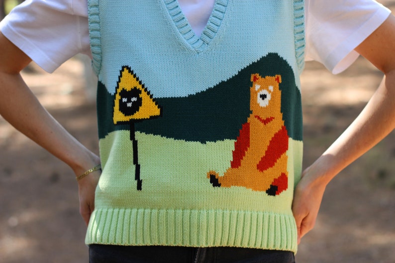 May include: A light blue, green, and yellow knitted vest with a bear and a warning sign graphic. The vest has a V-neck and a ribbed bottom.