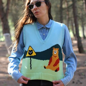 May include: A light blue knitted vest with a green, yellow, and brown bear and a yellow triangle with a black cat graphic. The vest has a V-neck and is worn over a light blue button-down shirt.