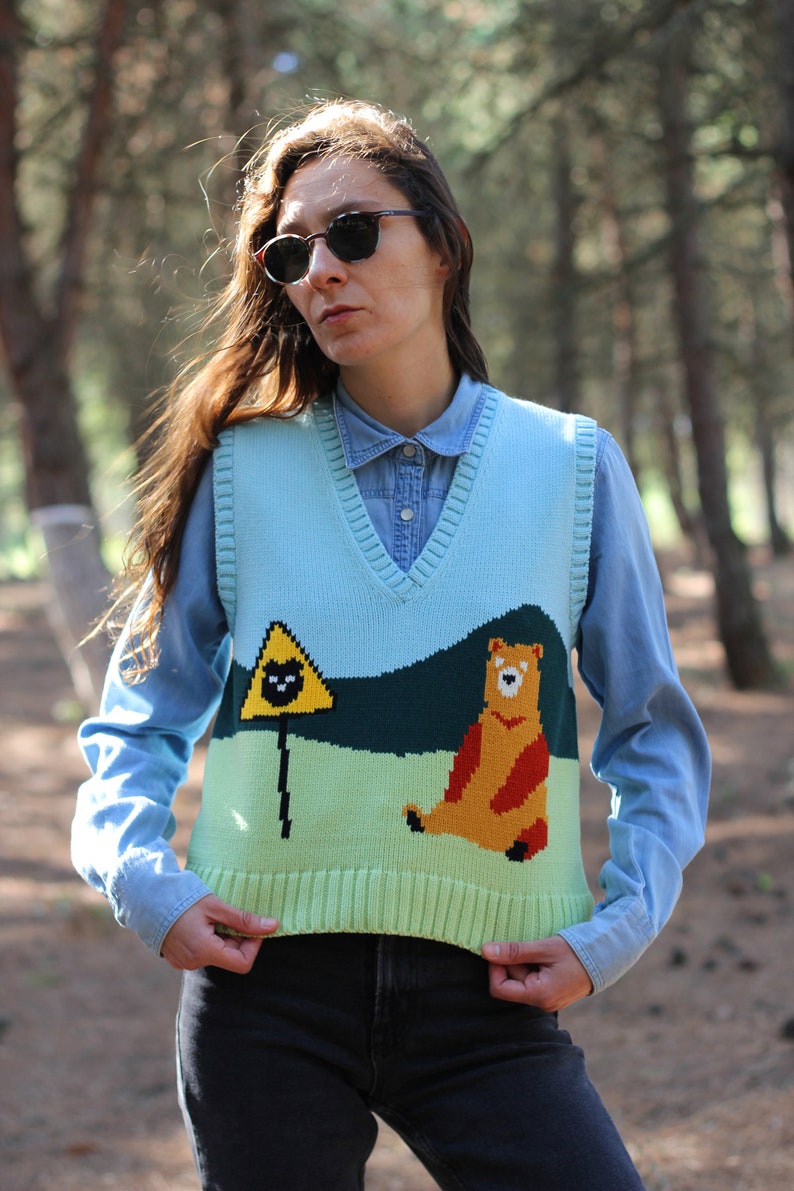 May include: A light blue knitted vest with a green, yellow, and brown bear and a yellow triangle with a black cat graphic. The vest has a V-neck and is worn over a light blue button-down shirt.