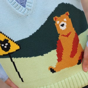May include: A light blue knitted vest with a brown bear and a yellow triangle with a black cat graphic. The vest has a green, light green, and dark green striped pattern.