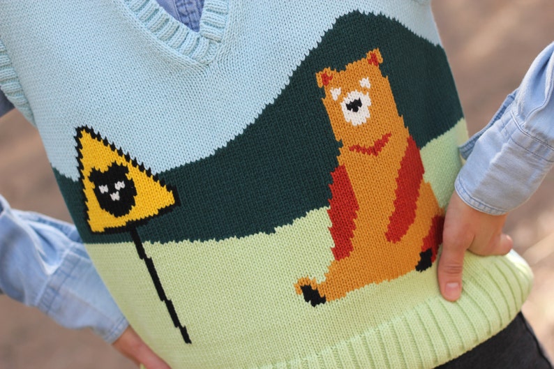 May include: A light blue knitted vest with a brown bear and a yellow triangle with a black cat graphic. The vest has a green, light green, and dark green striped pattern.