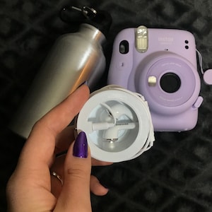Puede incluir: Una caja blanca con un par de auriculares blancos en su interior. La caja tiene la palabra "SAMSUNG" impresa en ella. La caja está siendo sostenida por una mano con esmalte de uñas morado. Una botella de agua plateada y una cámara Instax Mini morada están en el fondo.